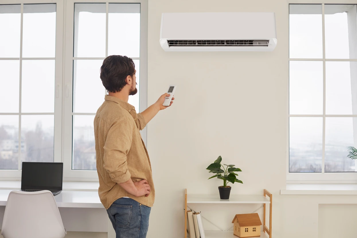 Homme d'affaires en chemise beige utilisant une télécommande pour contrôler un climatiseur mural dans un bureau moderne avec baies vitrées