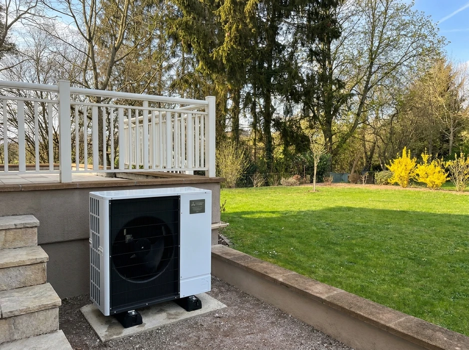 Unit&eacute; ext&eacute;rieure de pompe &agrave; chaleur air/eau install&eacute;e sur dalle b&eacute;ton, adoss&eacute;e &agrave; un mur de terrasse sur&eacute;lev&eacute;e avec garde-corps blanc